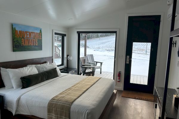 A cozy bedroom with a neatly made bed, a "Adventure Awaits" sign, and a window view of a snowy landscape.