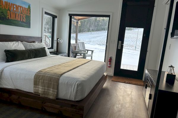 A cozy bedroom with a large bed, artwork on the wall, view outside, and a door leading to a snowy landscape.