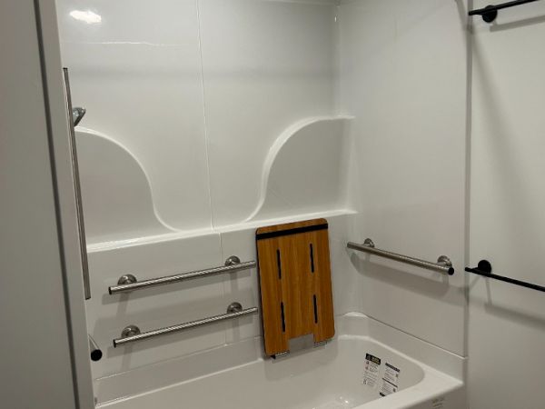 A bathroom with a white bathtub, safety grab bars, and a fold-down wooden seat. The floor is wood-look, and there's overhead lighting.
