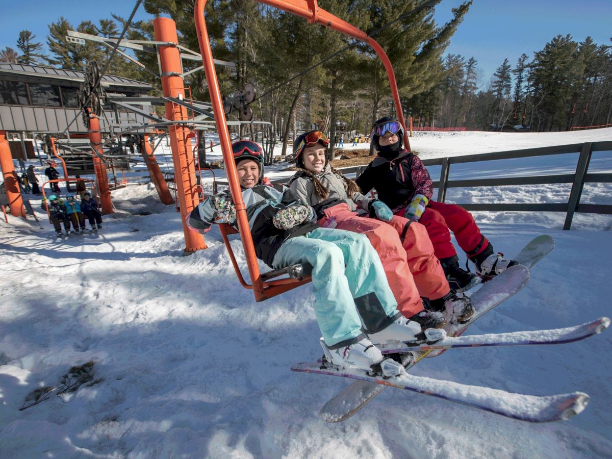 Three smiling skiers in colorful winter gear ride a bright orange ski lift together on a sunny day, with snow-covered ground, trees, and other skiers in the background.