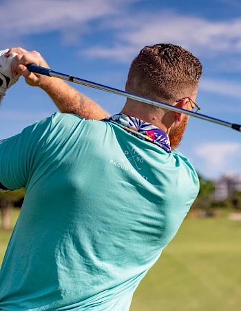 A person is swinging a golf club on a grassy course, wearing a turquoise shirt and gloves, with a clear blue sky in the background.