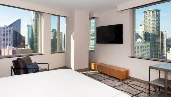 A modern hotel room with large windows, city views, a wall-mounted TV, a chair, and a bed. The decor is minimalistic and contemporary.