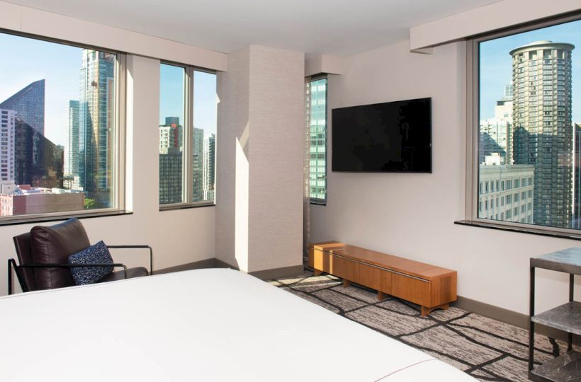 A modern hotel room with large windows, city views, a wall-mounted TV, a chair, and a bed. The decor is minimalistic and contemporary.