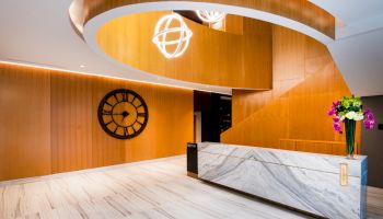 A modern lobby with a marble reception desk, wooden wall panels, a large clock, and hanging light fixtures with a spiral staircase overhead.