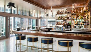 The image features a modern, well-lit bar with high stools and a large variety of bottles on shelves, overlooking a city through large windows.