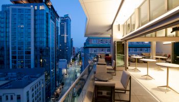 The image shows a modern cityscape view from a high-rise balcony at dusk, with chairs and tables outside and glass buildings lining the street.