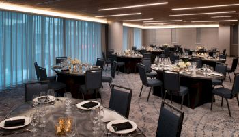 The image shows an elegantly set dining hall with round tables, black chairs, candles, and tableware, likely ready for an event or banquet.
