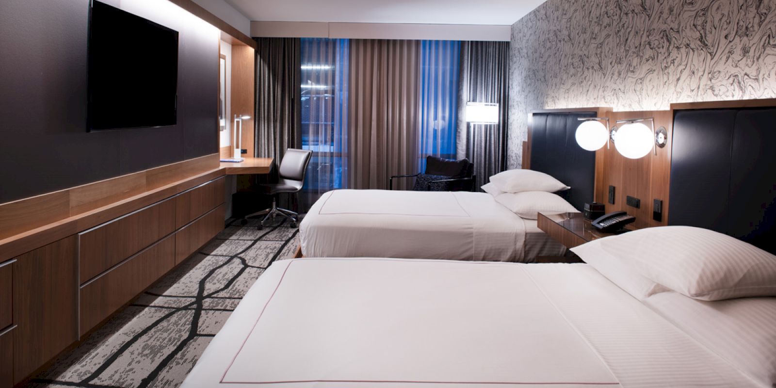 The image shows a modern hotel room with two neatly made beds, a desk, a flat-screen TV, and stylish decor including patterned wallpaper and contemporary lighting.