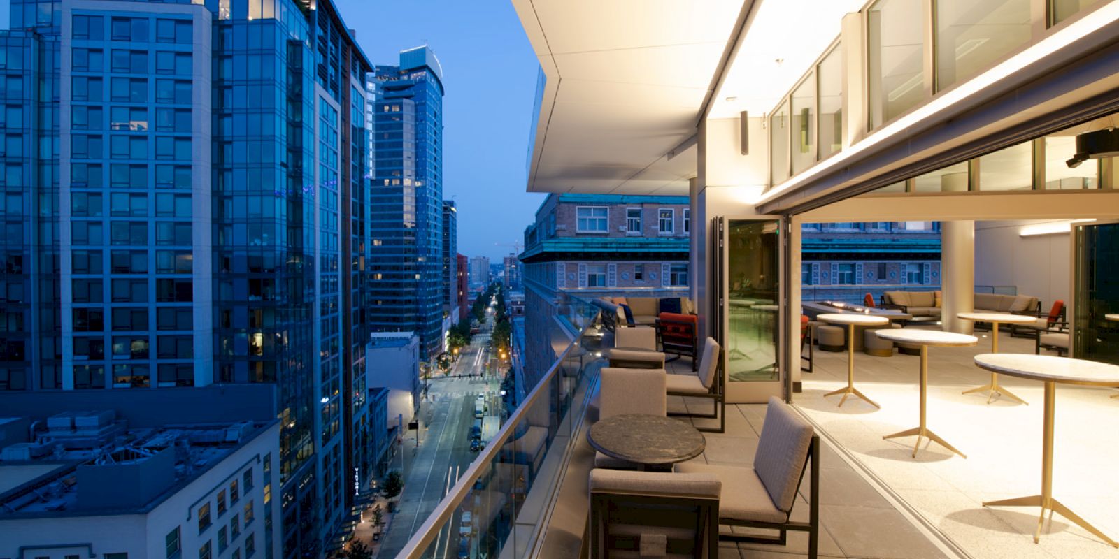 A rooftop terrace with chairs and tables, overlooking a street and surrounding modern buildings during twilight hours.