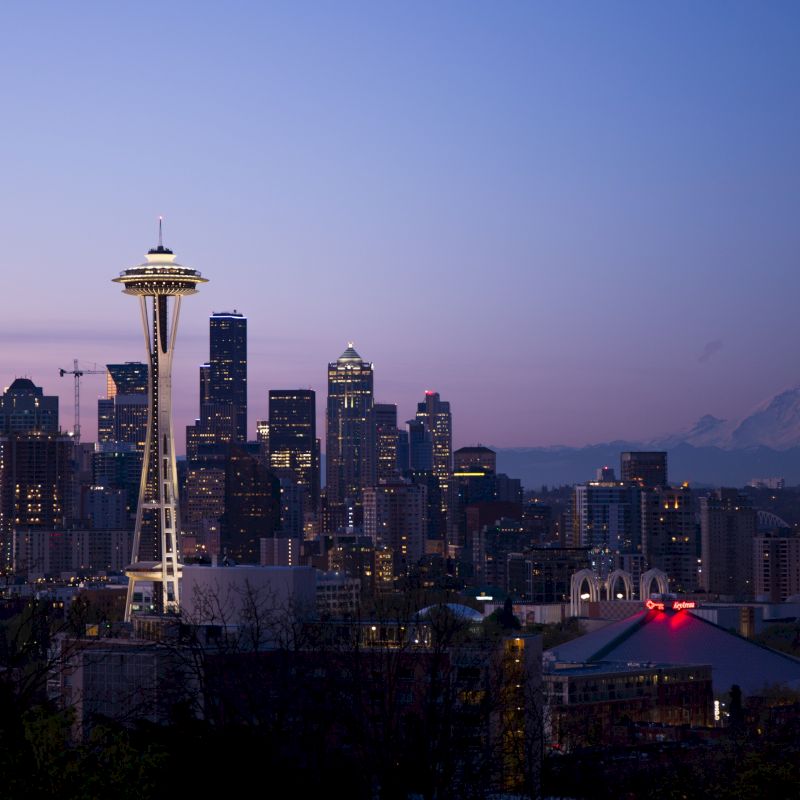 The image shows the Seattle skyline at dusk, featuring the Space Needle prominently with Mt. Rainier visible in the background under a twilight sky.