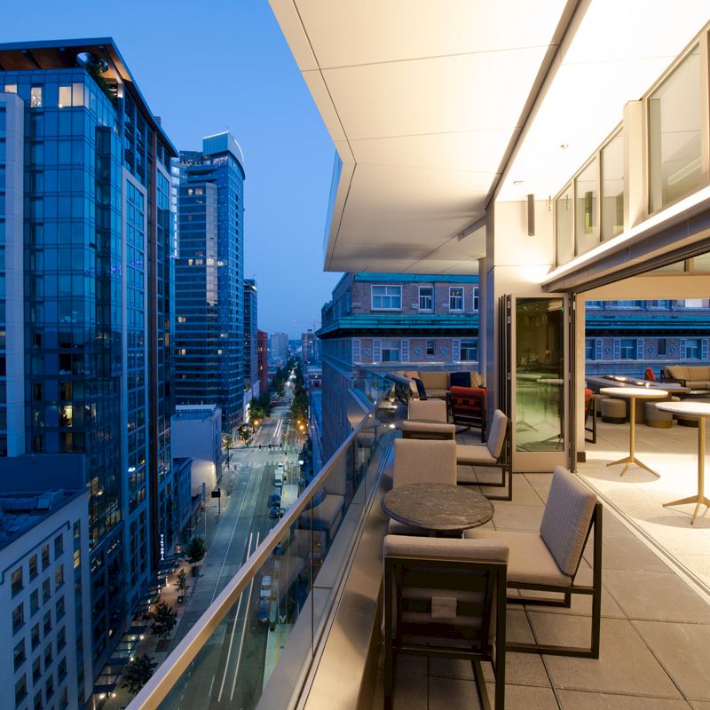 The image shows a modern cityscape at dusk with a high-rise view. There is an outdoor balcony with tables and chairs and buildings visible.