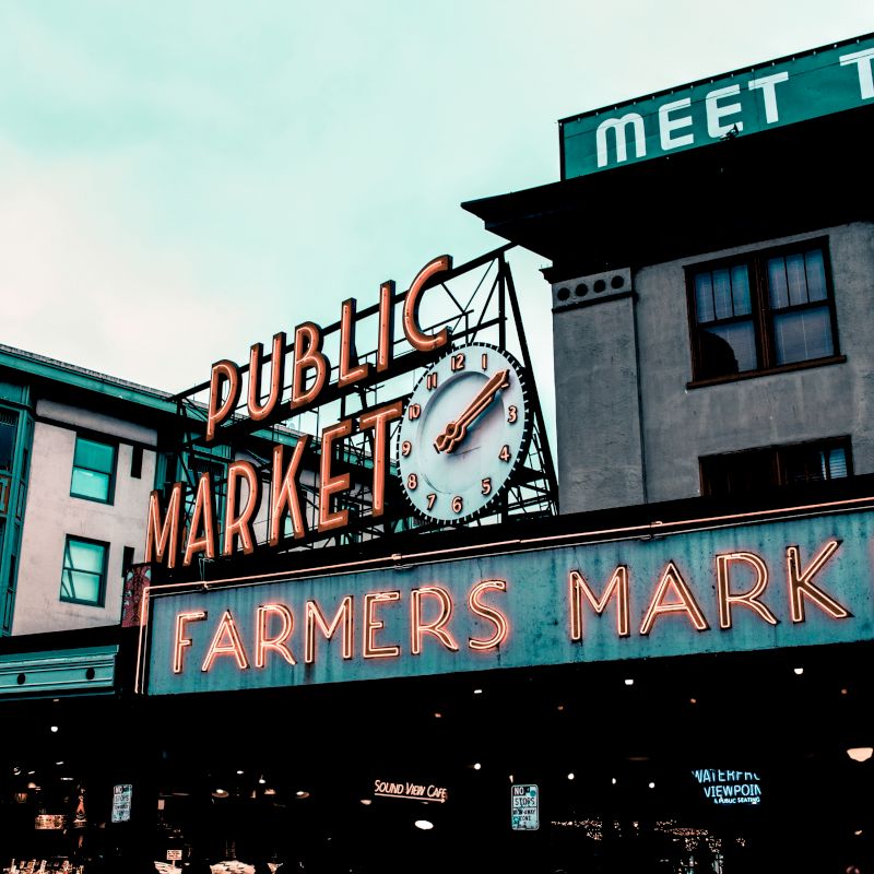 The image shows an exterior view of a classic public market sign with a clock and neon lights, advertising a farmers market in an urban setting.