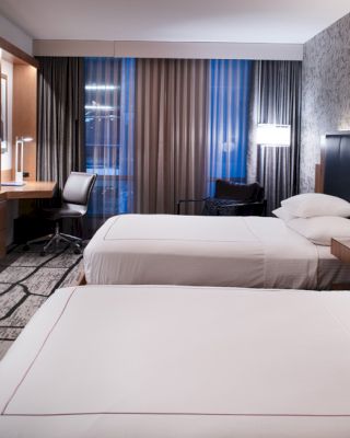 A modern hotel room features two neatly made beds, a flat-screen TV, a desk with a chair, and contemporary decor with patterned walls and curtains.