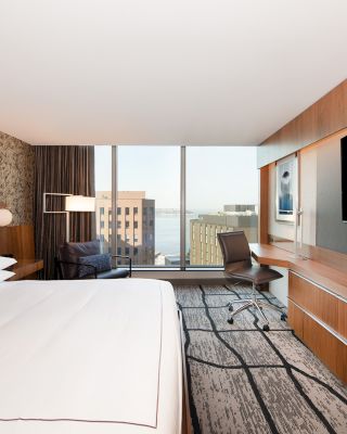 This image shows a modern hotel room with a king-sized bed, a large TV, a work desk, and a window with a city view.