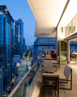 A modern balcony at twilight, featuring tables and chairs, overlooks a city street lined with high-rise buildings.