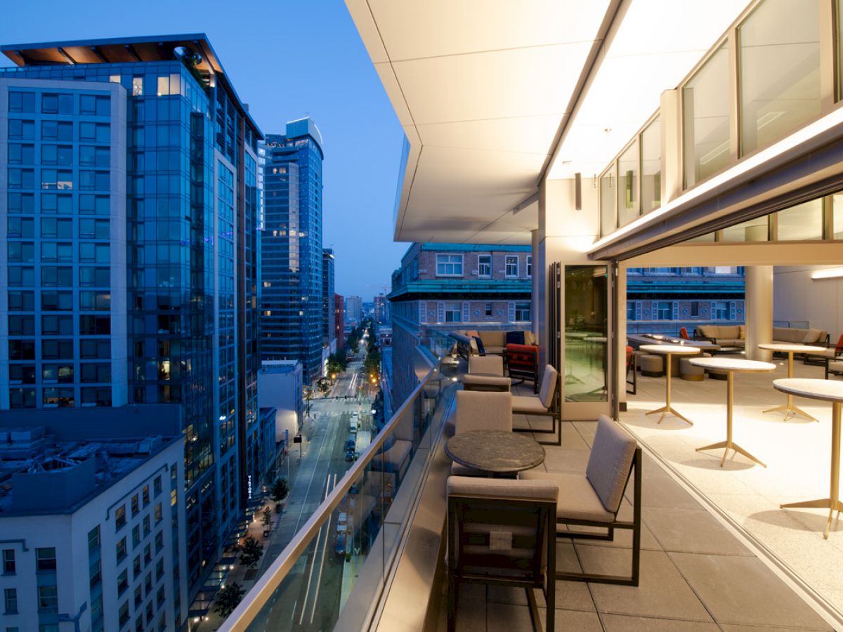 The image shows a modern balcony with seating and tables, overlooking a cityscape with tall buildings during the evening.