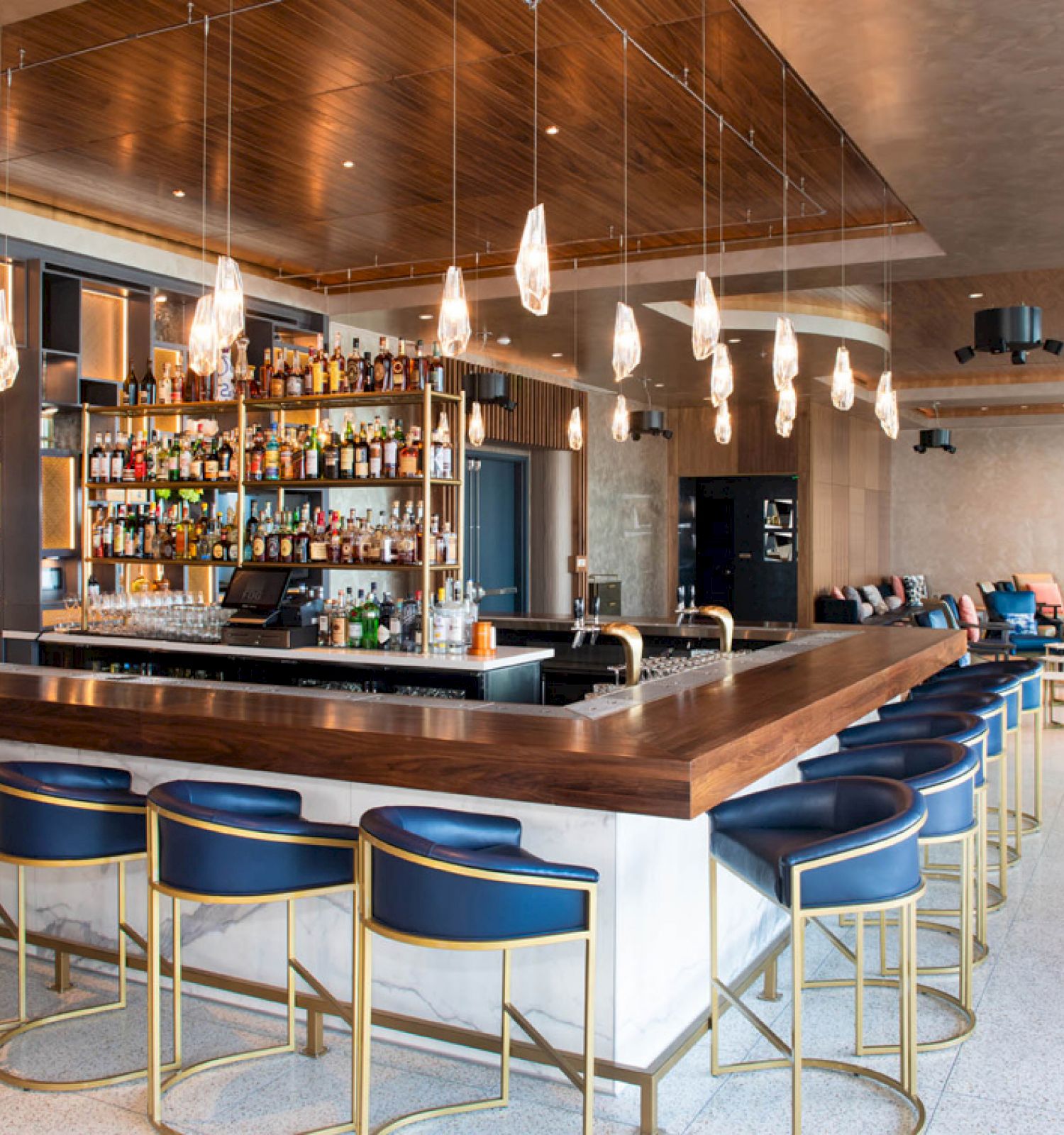 This image shows a modern bar with a long counter, stylish bar stools, various hanging lights, and shelves stocked with alcohol bottles in the background.