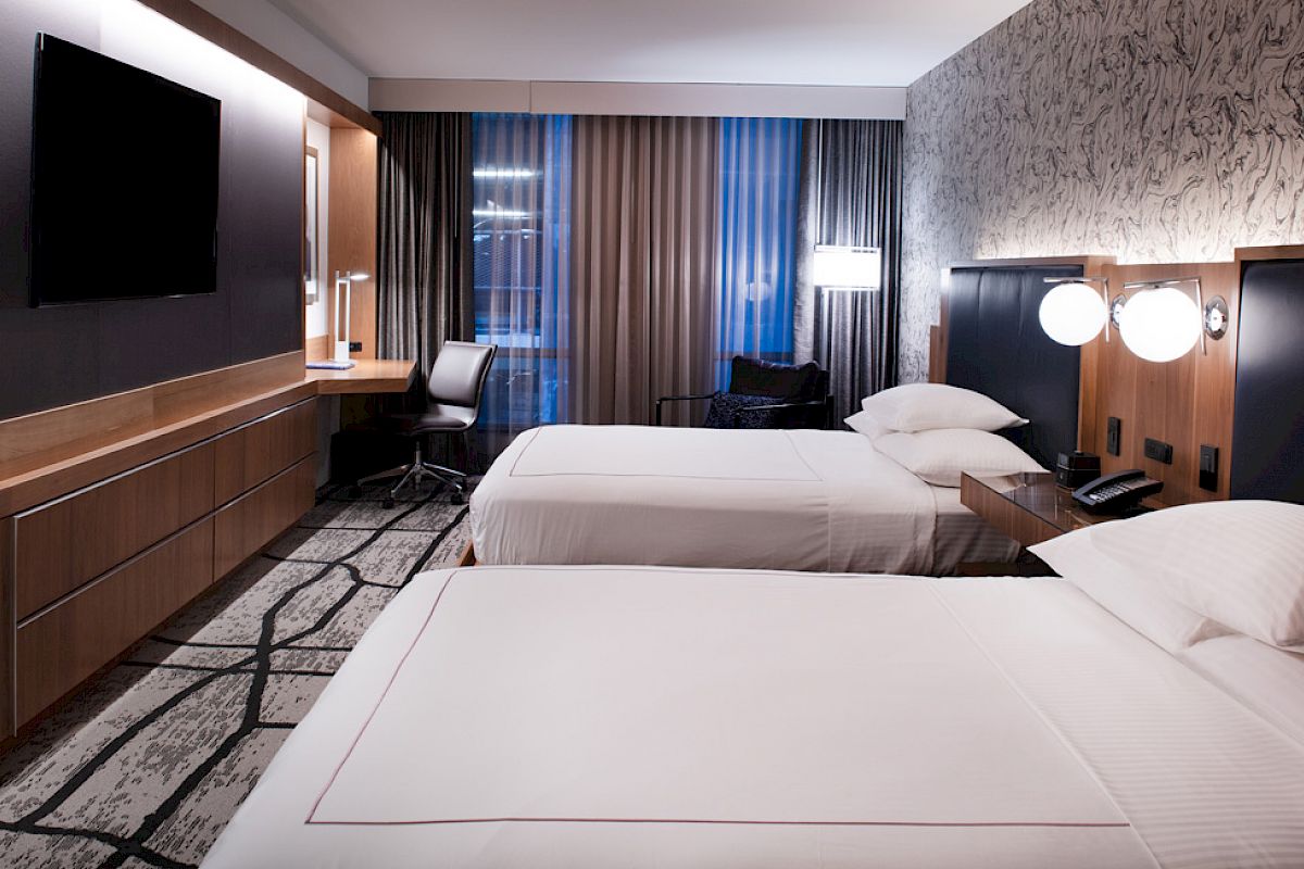 The image shows a modern hotel room with two beds, a TV, a desk with a chair, bedside lamps, and patterned wallpaper.