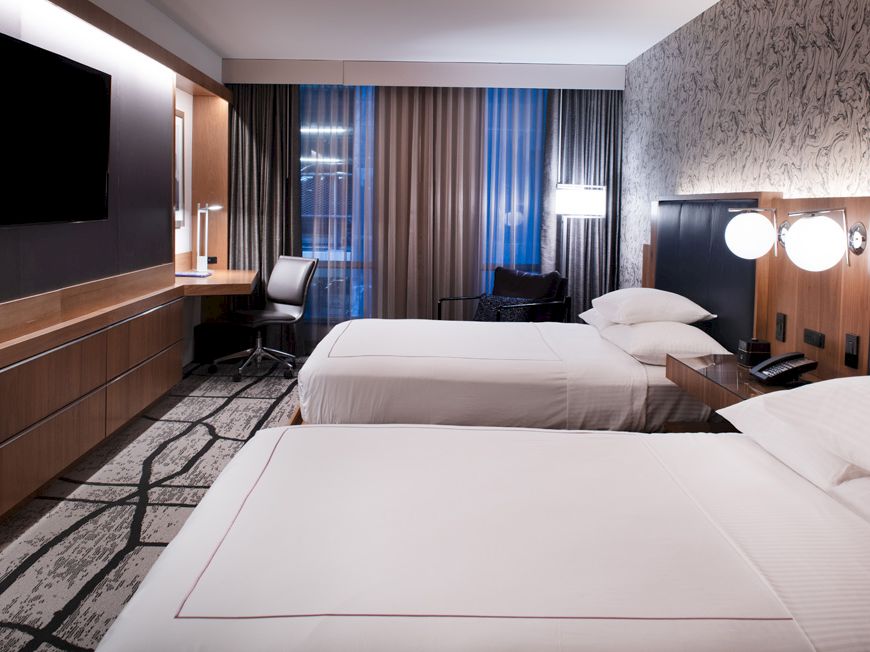 The image shows a modern hotel room with two beds, a TV, a desk with a chair, bedside lamps, and patterned wallpaper.