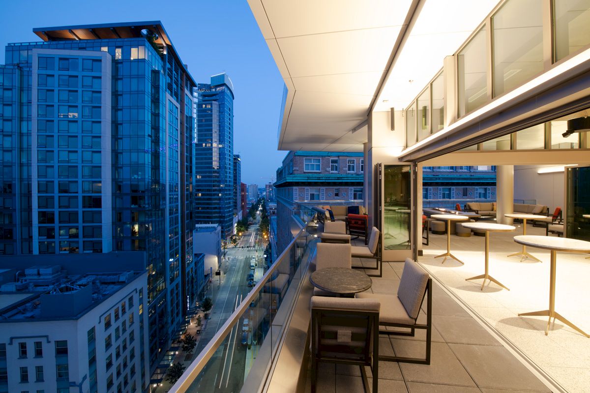 A modern urban balcony at dusk, overlooking a city street with tall buildings. The balcony features chairs, tables, and indoor-outdoor space.