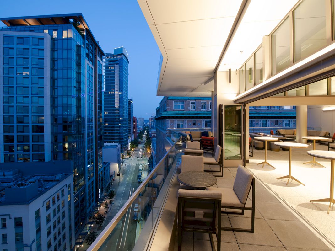A modern urban balcony at dusk, overlooking a city street with tall buildings. The balcony features chairs, tables, and indoor-outdoor space.