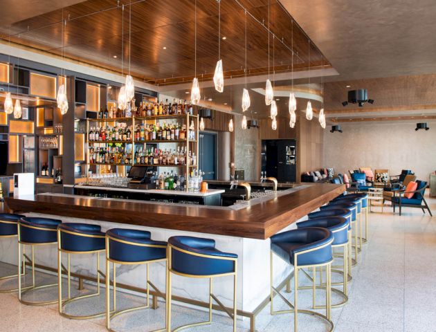 A modern bar with a well-stocked shelf and elegant seating area, featuring blue barstools and pendant lights, creating a cozy atmosphere.