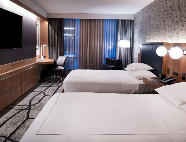 The image shows a modern hotel room with two neatly made beds, a desk and chair by the window, a wall-mounted TV, and stylish decor.