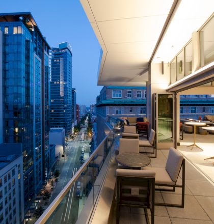 This image features a modern balcony with outdoor seating overlooking a cityscape with tall buildings and a street below during evening time.