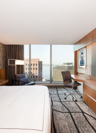 A modern hotel room with a large bed, wall-mounted TV, desk, chair, and floor-to-ceiling windows offering a city view and natural light.