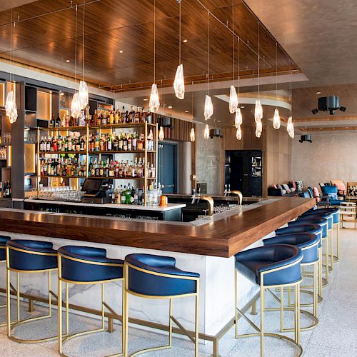 A modern bar with a well-stocked shelf of bottles, elegant lighting, blue bar stools, and additional seating areas in the background.