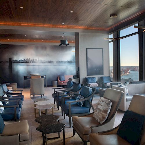 This image shows a modern, comfortable lounge area with plush seating, large windows offering a scenic view of the water and a cityscape mural.