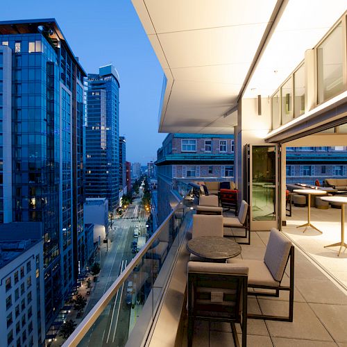 A modern balcony overlooking a cityscape at dusk, featuring glass railings, outdoor seating, and tall buildings lining the street below.