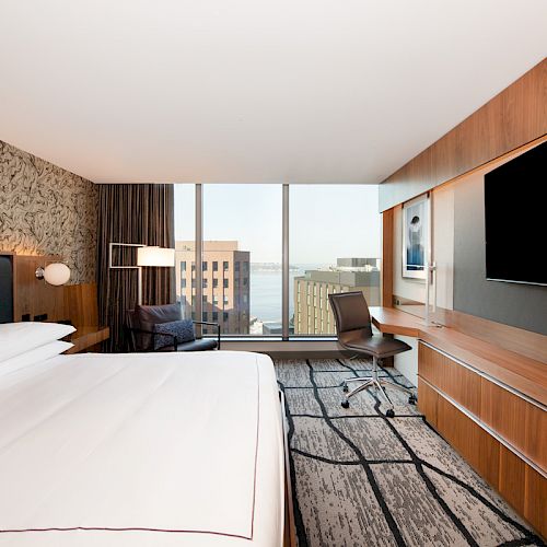A modern hotel room with a large bed, wall-mounted TV, desk, chair, and window overlooking a cityscape with buildings and water in the distance.