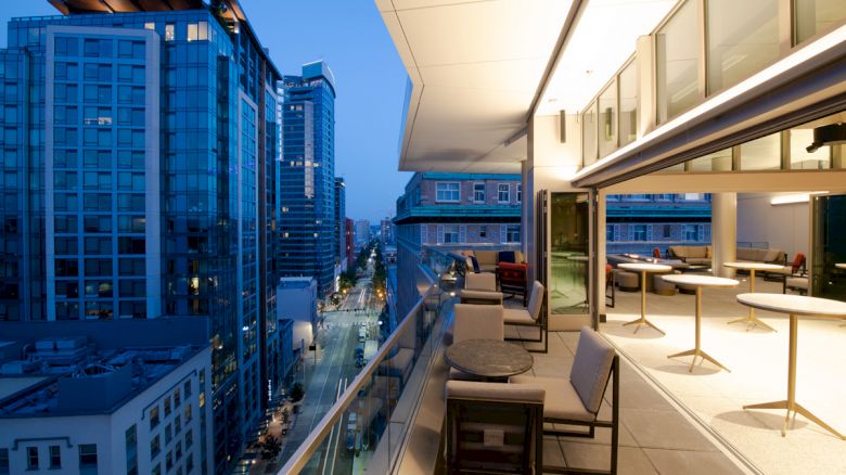 The image depicts a modern balcony overlooking a cityscape with tall buildings. The balcony has seating and tables, illuminated in evening light.