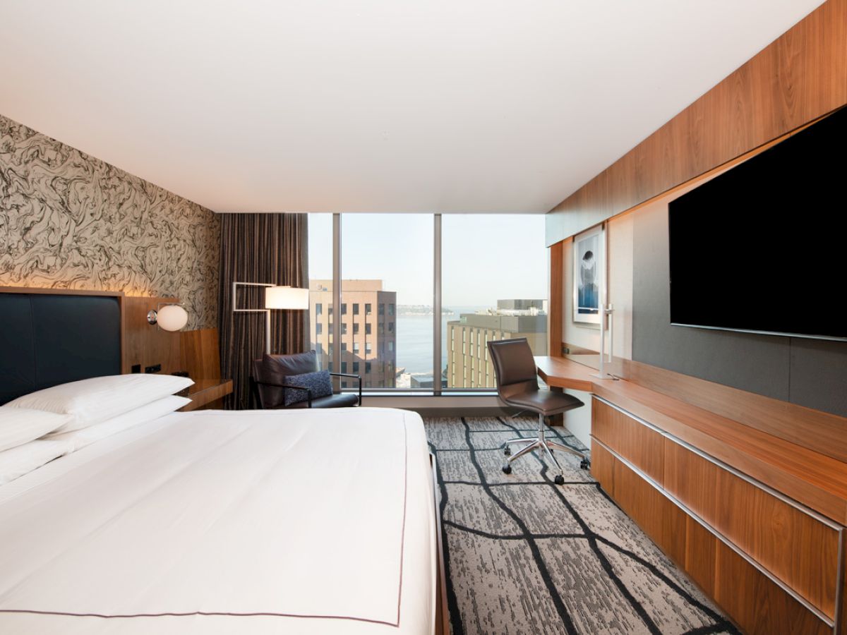 A modern hotel room with a king-size bed, large flat-screen TV, desk, and a view of city buildings through floor-to-ceiling windows.