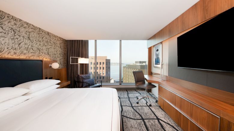 A modern hotel room with a king-size bed, large flat-screen TV, desk, and a view of city buildings through floor-to-ceiling windows.