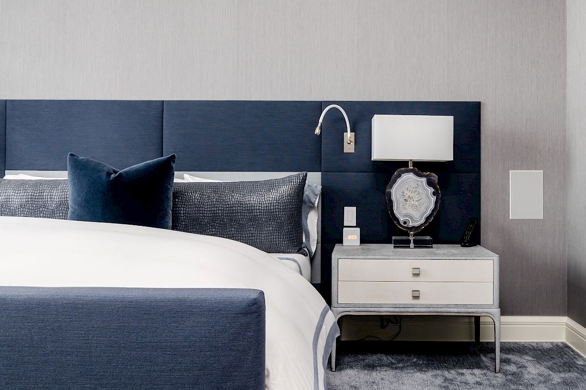 A modern bedroom features a navy blue and white theme, with a bed, nightstand, lamp, and a decorative item.