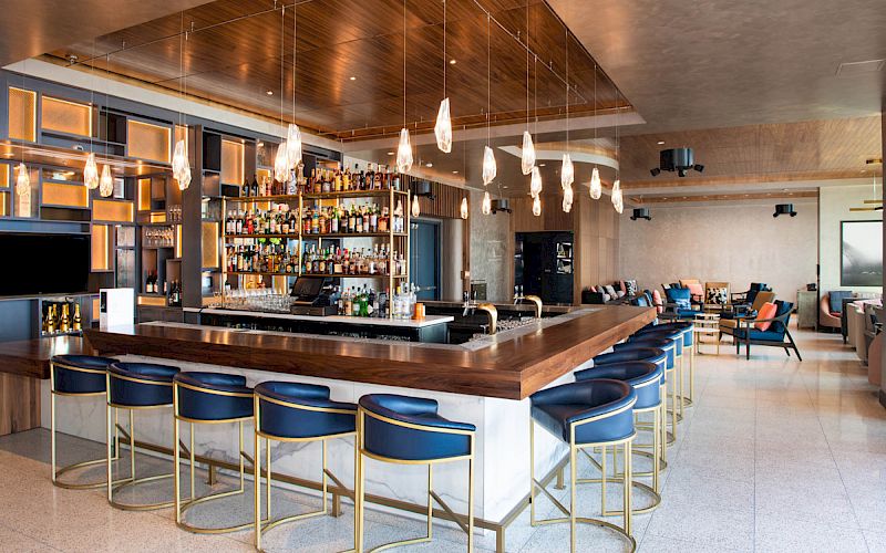 A modern bar features a long counter with blue stools, a large selection of bottles on shelves, hanging pendant lights, and a stylish lounge area.
